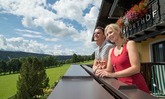 A couple stands on a balcony and enjoys the view of the green landscape. The sky is clear with some clouds, and flowers beautify the balcony.