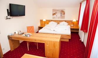 A cozy hotel room with a double bed and a wooden table. The walls are adorned with red curtains and there is a television on the wall.