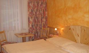 A cozy bedroom with a double bed and warm colors. Next to the bed is a table with chairs and a window with curtains.