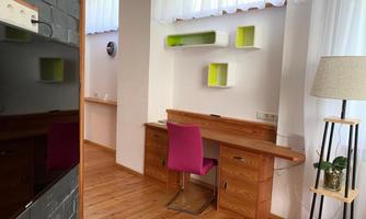 A modern workspace with a wooden desk and a pink chair. On the wall, there are colorful shelves and a bright window that lets in a lot of light.