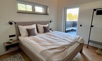 A modern bedroom with a large bed and bright wood details. The room has large windows that let in plenty of daylight.