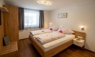 A cozy bedroom with a double bed and floral bedding. The walls are light, and there is a window with curtains as well as wooden shelves.