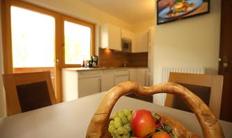 A cozy kitchen area with a wooden table and a fruit basket. In the background, you can see the modern kitchen and a television.