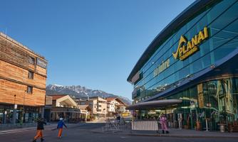 A modern entrance area of a ski resort with large windows and the inscription "Planai". In the background, mountains and a clear blue sky can be seen.