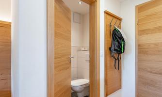 A bright hallway with open access to a toilet. A backpack hangs on the wall, and the doors are made of wood.