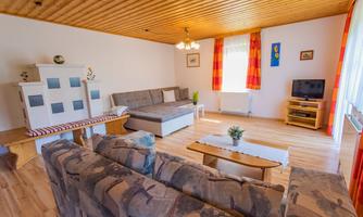 A cozy living room with a wooden ceiling and light parquet flooring. There is a sofa, a television, and large windows with orange curtains.