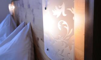 A modern wall lamp with floral patterns softly illuminates above a wooden bed. Beneath it lie two white pillows.