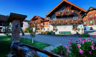 A cozy alpine building with blooming balconies and a fountain in the foreground. The garden is decorated with colorful flowers and the sky is clear and blue.