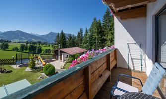 A beautiful balcony with a view of the mountains and a green landscape. Flowers are blooming and a pool can be seen in the garden.