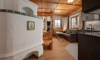 A cozy living space with a traditional tiled stove and wooden ceiling. The decor is modern and inviting, with a seating area and a dining table.