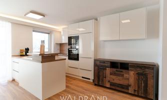 A modern kitchen with light wooden flooring and white glossy furniture. On the left side, there is a kitchen island, and on the right side, there is a dark wooden cabinet.