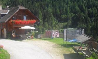 A picturesque farmhouse with blooming balcony flowers and a shade-providing umbrella. In the background, a clothes drying rack and a trampoline can be seen on a green lawn.