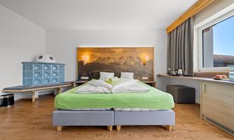 A modern bedroom with a large bed and a bright wooden floor. There is a desk by the window and an elegant blue seating area.