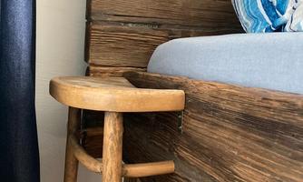A rustic bed with a wooden finish and a gray mattress. Next to the bed is a simple, round wooden chair.