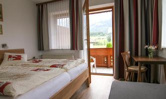 A cozy bedroom with two beds and a small table. The door leads to a balcony with a view of the mountains.