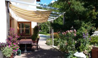 An inviting outdoor area with a table and chairs under a sunshade. Colorful flowers and green plants surround the space, creating a pleasant atmosphere.