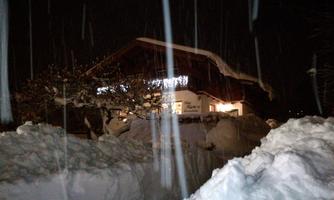 A cozy house in a snowy landscape at night. The snow is deep, and it is lightly snowing.