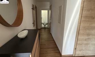 A bright hallway with wooden flooring and modern furniture. On the left, there is a dresser with a round mirror.