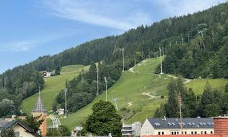 A green hilly landscape with ski lifts and dense forests. In the foreground, houses and trees can be seen.