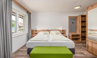 A modern bedroom with a large double bed and a green bench. The room has bright walls and plenty of natural light.
