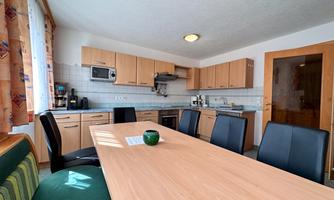 A bright kitchen with wooden cabinets and a large table. The room is modernly furnished and has windows with curtains.