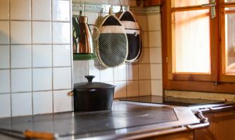 A cozy kitchen with a stove and a black pan. Cooking utensils hang on the wall and a window lets in light.