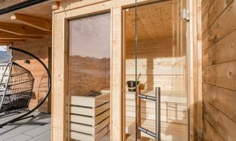 A modern wooden sauna with large glass windows. In the background, a cozy rocking chair can be seen.