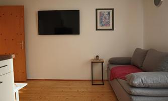 A cozy living room with a gray sofa and a TV on the wall. A framed picture hangs on the wall, and a lamp is on a small table.