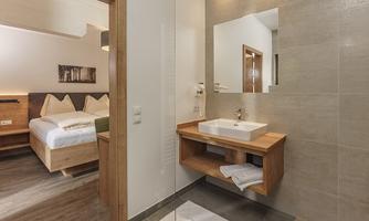 A modern bathroom with a large shower and an elegant sink. In the background, a cozy bedroom is visible.