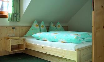 A cozy bedroom with a wooden bed and colorful cushions. The walls are light, and the room has a friendly atmosphere.