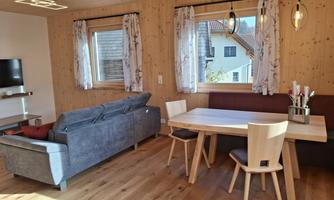 A cozy living room corner with a gray sofa and a wooden dining area. Large windows let in plenty of daylight and offer a view outside.