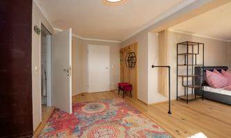 A bright entrance area with wooden flooring and a colorful rug. There is an open door and a modern shelf visible.