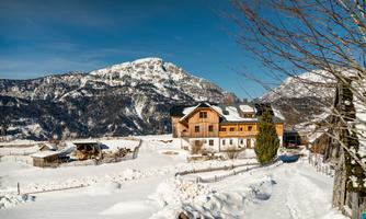 A picturesque winter landscape with snow-covered hills and mountains in the background. In the foreground stands a traditional wooden house surrounded by white snow.
