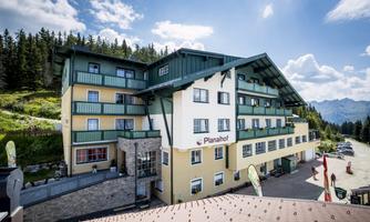A modern hotel with several floors, surrounded by trees and mountains. The clear sky and sunny surroundings create an inviting atmosphere.
