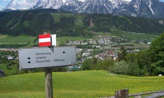 A signpost points in different directions with the names of places in the Alps. In the background, impressive mountains and a green hilly landscape can be seen.
