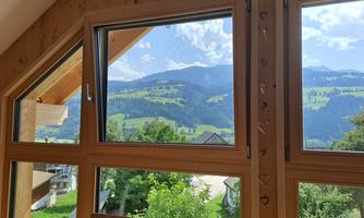 A view from a wooden window onto a green landscape with hills and mountains. The sky is blue with some clouds.