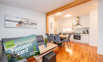 A modern living room with wooden floors, a cozy sofa, and a dining area. In the foreground, there is a summer tour map that provides information about local activities.