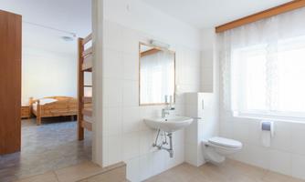A bright bathroom with a sink, toilet, and a large mirror. In the background, wooden beds are visible.