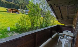 A balcony with a view of a green meadow and trees. In the background, wooden buildings and mountains can be seen.