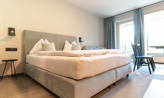 A modern hotel room with a large, comfortable bed and two chairs. Large windows let in plenty of daylight.