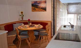 A cozy kitchen corner with a round table and a banquette. The table is set with dishes and flowers, while an open kitchen counter is visible in the background.