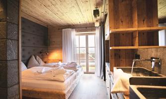 A cozy room with wooden ceilings and modern furniture. There is a large bed, a window front, and an open kitchenette.