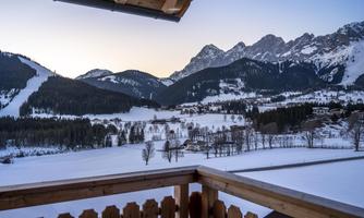 A winter mountain landscape with snow-covered hills and majestic mountains in the background. The view includes a tranquil, snowy setting with individual trees.