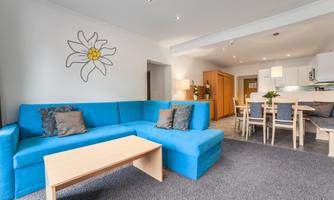 A modern living room with a blue couch and a wooden table. In the background, there is a dining area with a table and chairs.