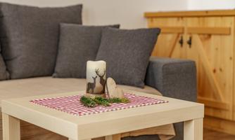 A cozy living room with a gray upholstered sofa and a wooden table. On the table, there is a decorative candle and a small wooden accent with some greenery.
