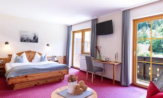 A cozy hotel room with wooden decor, a large bed, and a desk. The windows let in plenty of daylight and provide access to a balcony.