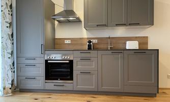A modern kitchen with gray furniture and wood details. It features a stove, a refrigerator, and a small coffee machine.
