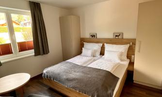 A cozy bedroom with a double bed and a table. The room has hardwood floors and large windows that provide plenty of light.