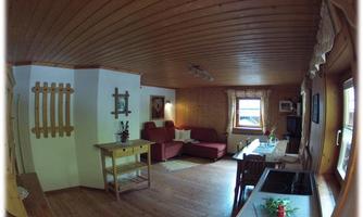 A cozy living room with wooden paneling, a red sofa, and a dining table. Light streams through the window and there is a small kitchen in the background.