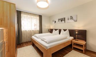 A modern bedroom with a double bed and light wooden furniture. The large windows provide plenty of daylight and a cozy atmosphere.